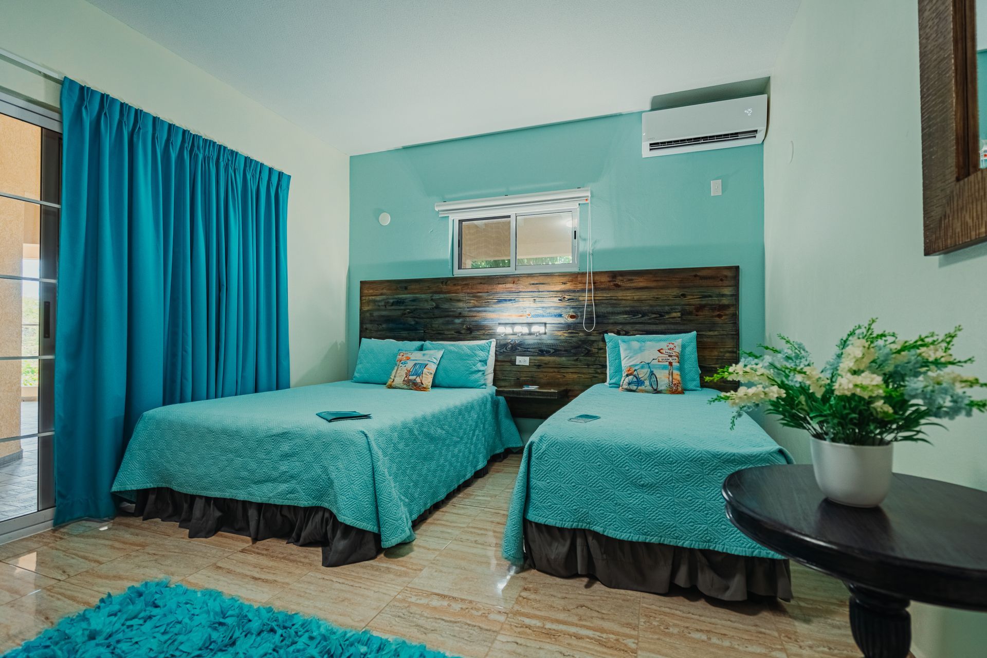 Turquoise twin-bed hotel room with teal curtains, wall art, and a small table with flowers