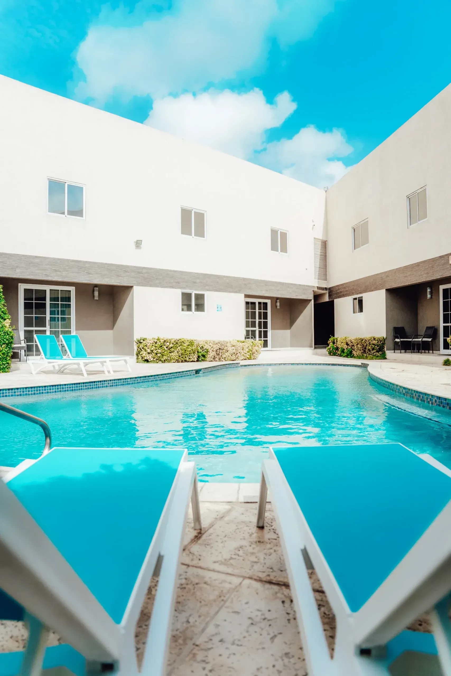 Bright courtyard pool with turquoise water, white buildings, and blue lounge chairs under a clear sky