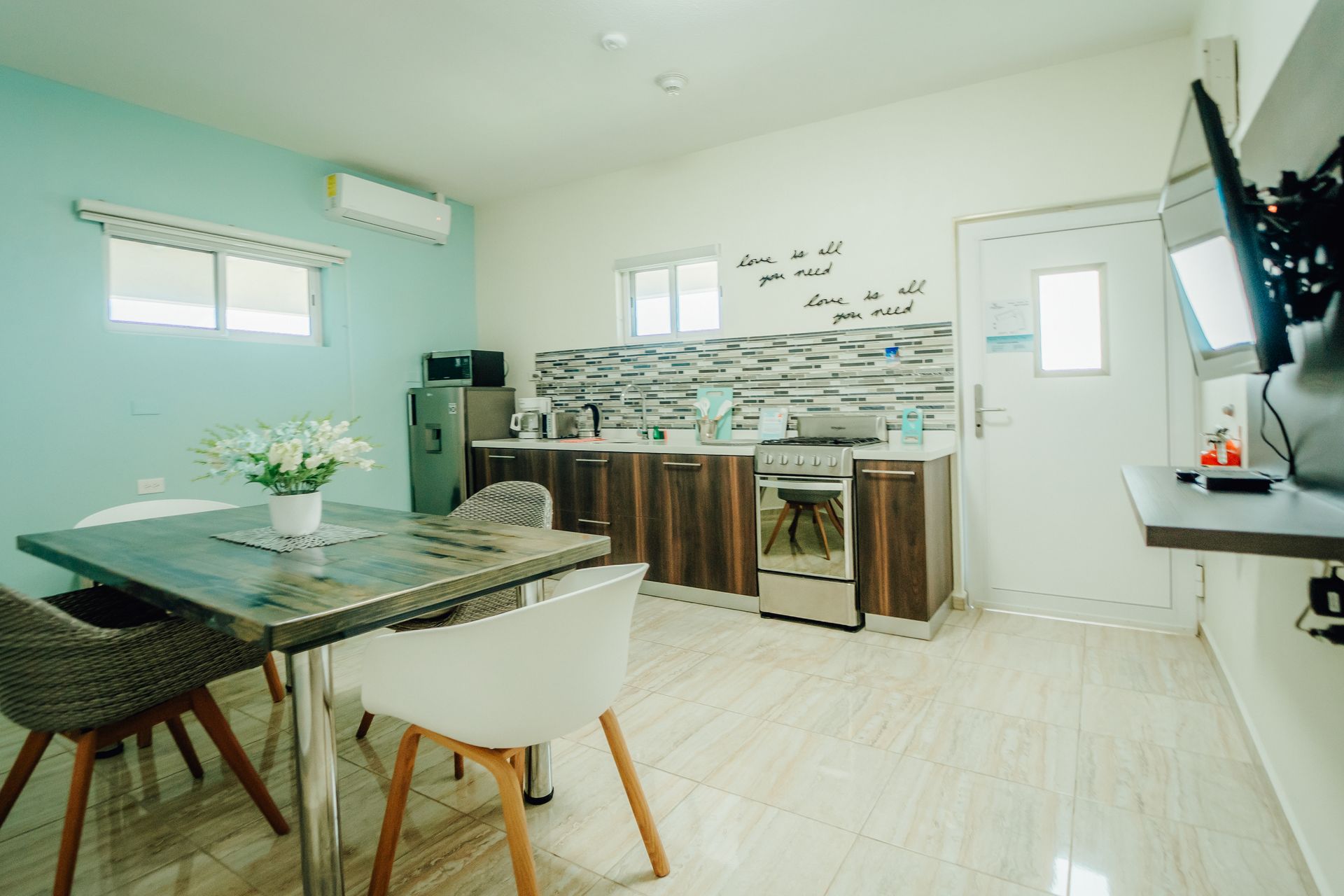 Bright dining area with glass table, white chairs, and a kitchen counter against pale green walls.
