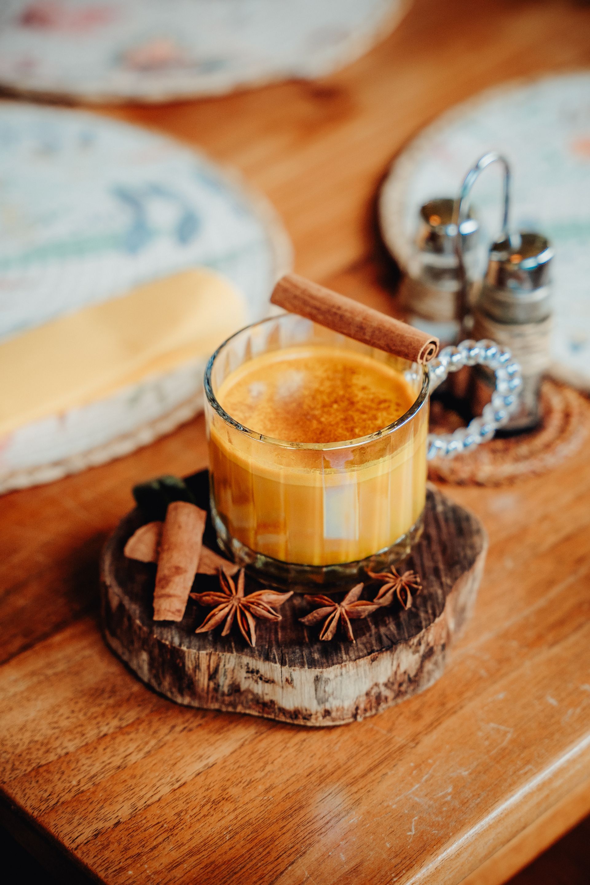 A glass of spiced latte on a wooden coaster with cinnamon sticks, star anise, and a metal spoon on a table.