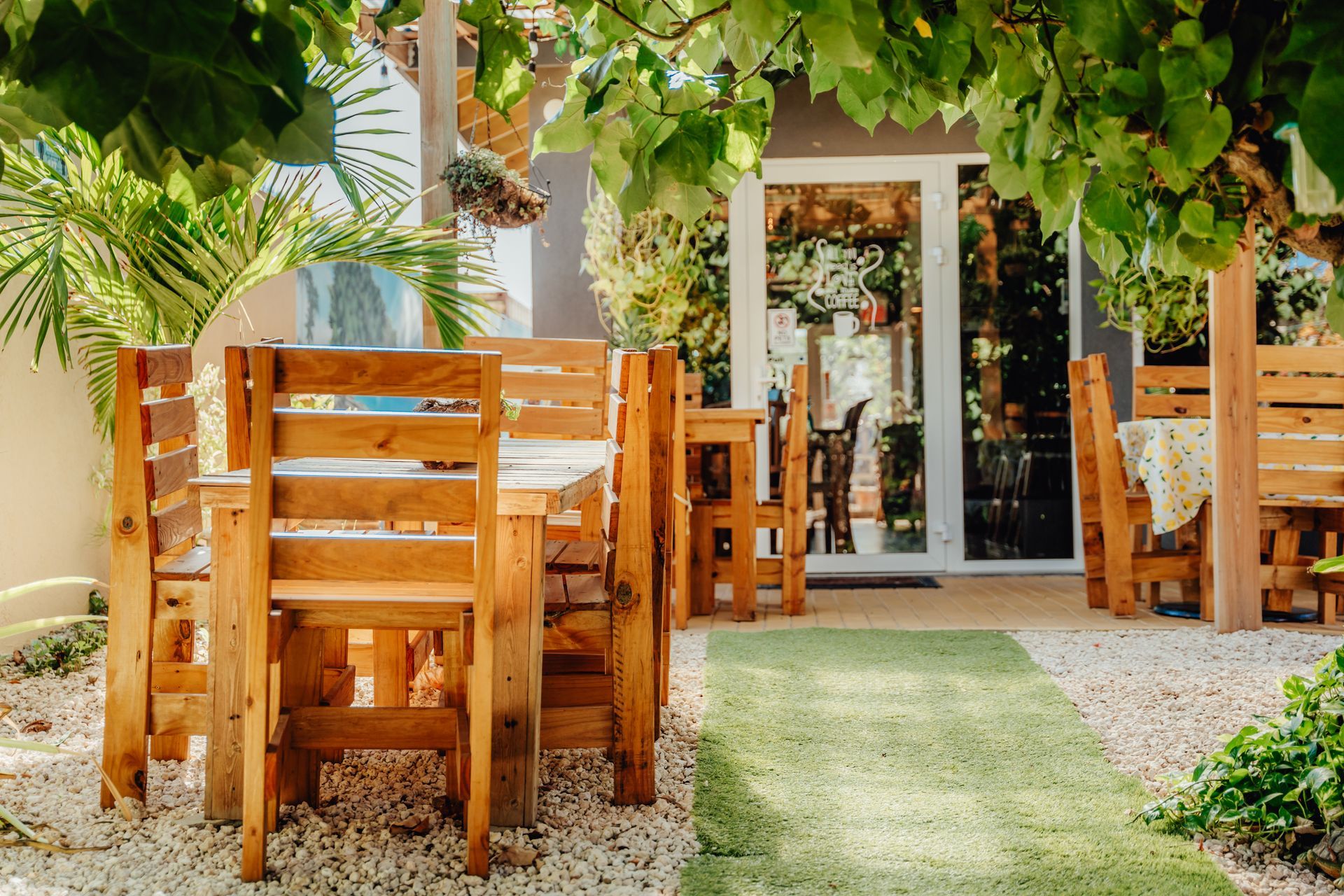 Outdoor café patio with wooden tables and chairs under leafy vines, leading to a glass door entrance