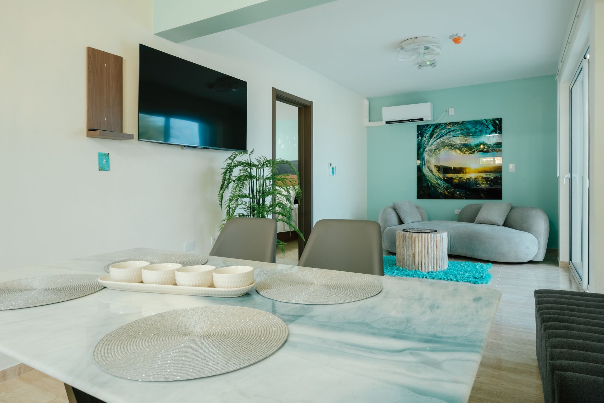 Bright modern apartment living room with white dining table, gray sofa, wall TV, and ocean artwork.
