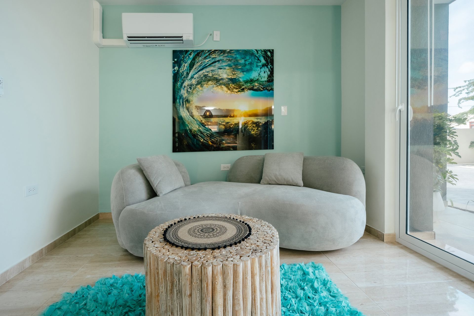Modern living room with a gray sofa, round wood table, turquoise rug, and wall art beneath an air conditioner