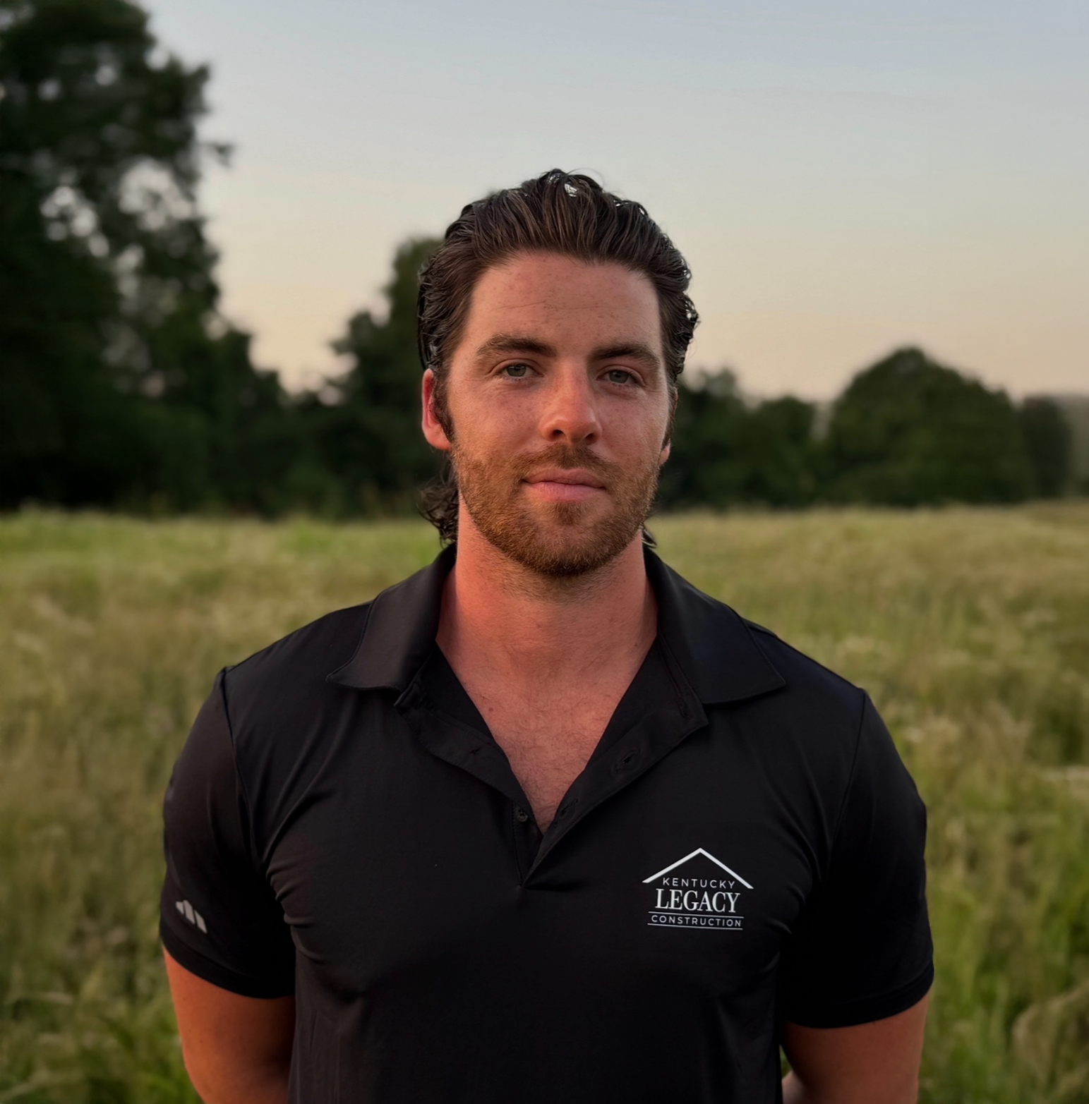 Man in black polo shirt standing outdoors at sunset with trees in the background
