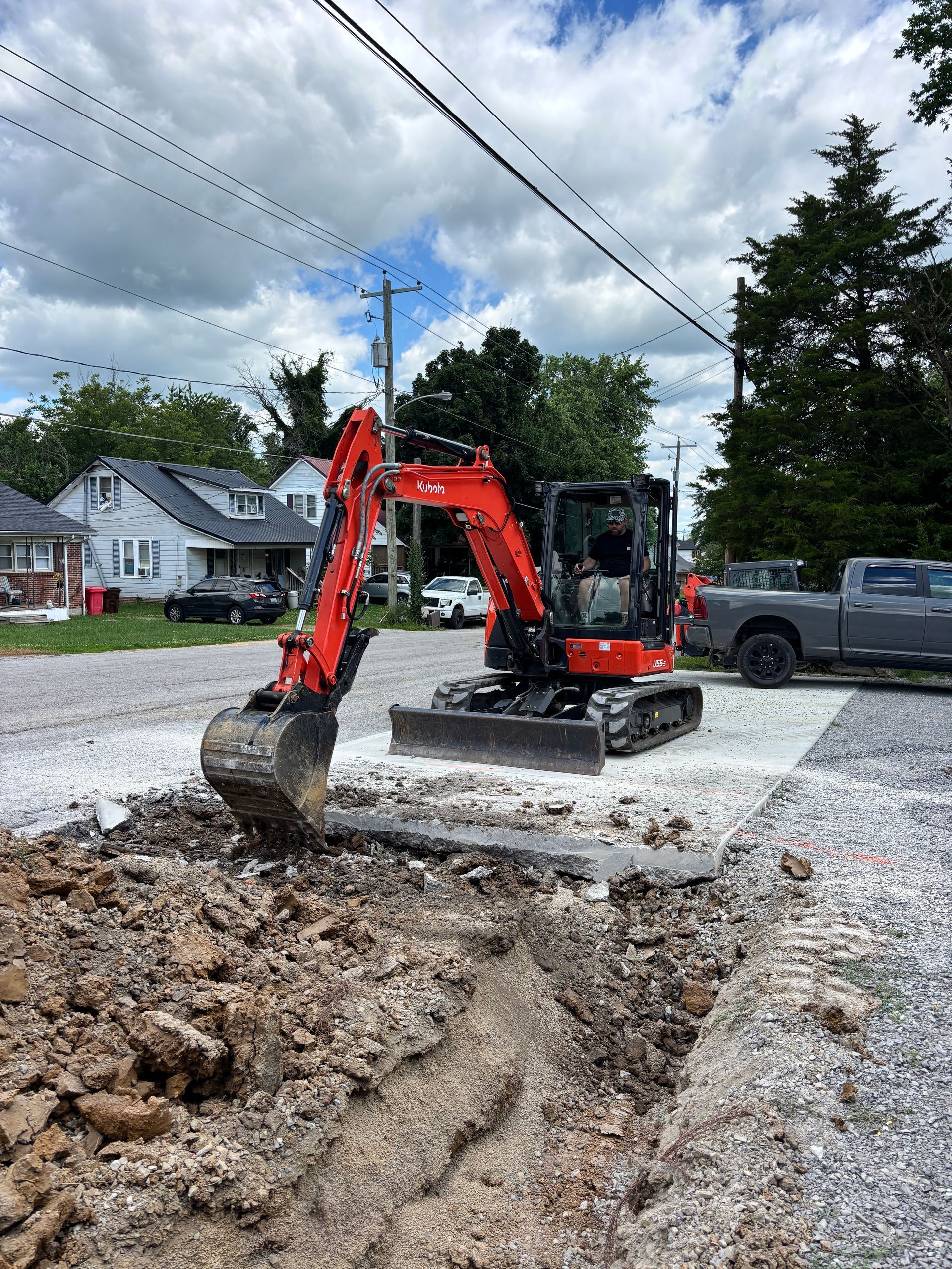 Orange excavator digging gravel on a residential street under a cloudy sky