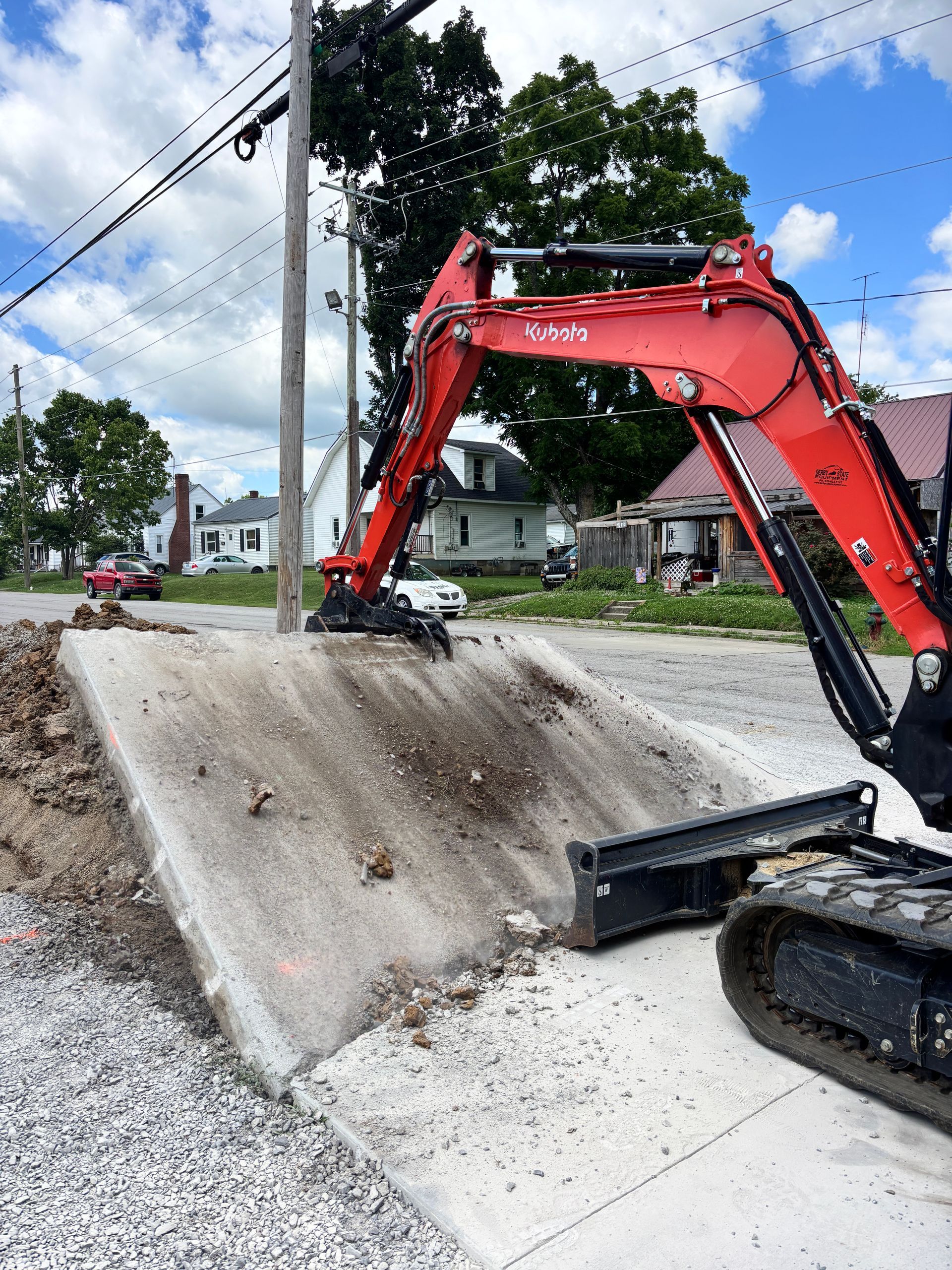Red excavator on a concrete roadside barrier, clearing debris beside a gravel street