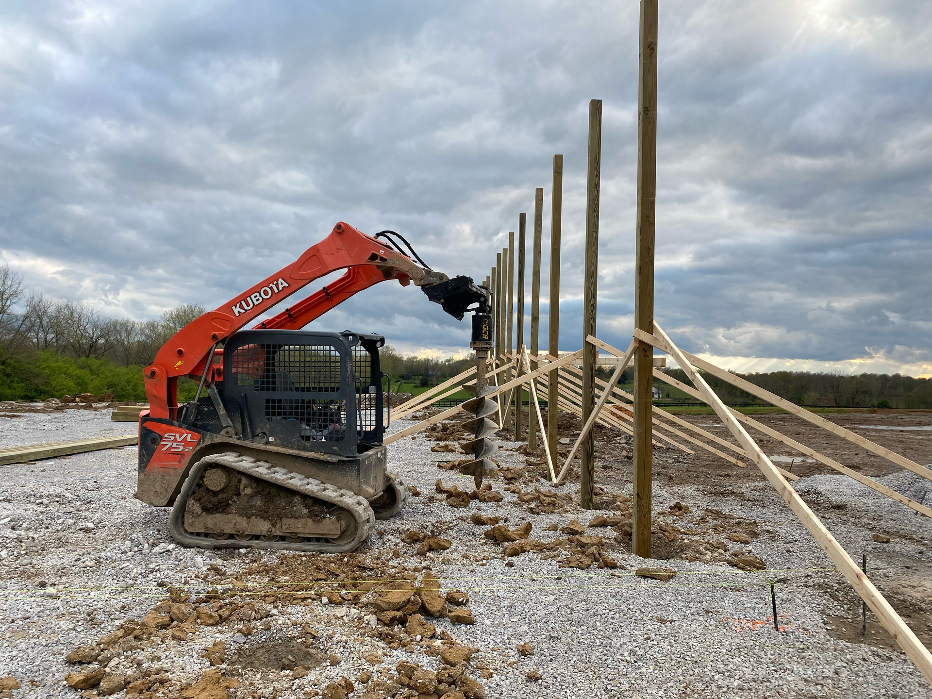 Mini excavator lifting wooden posts at a construction site under a cloudy sky