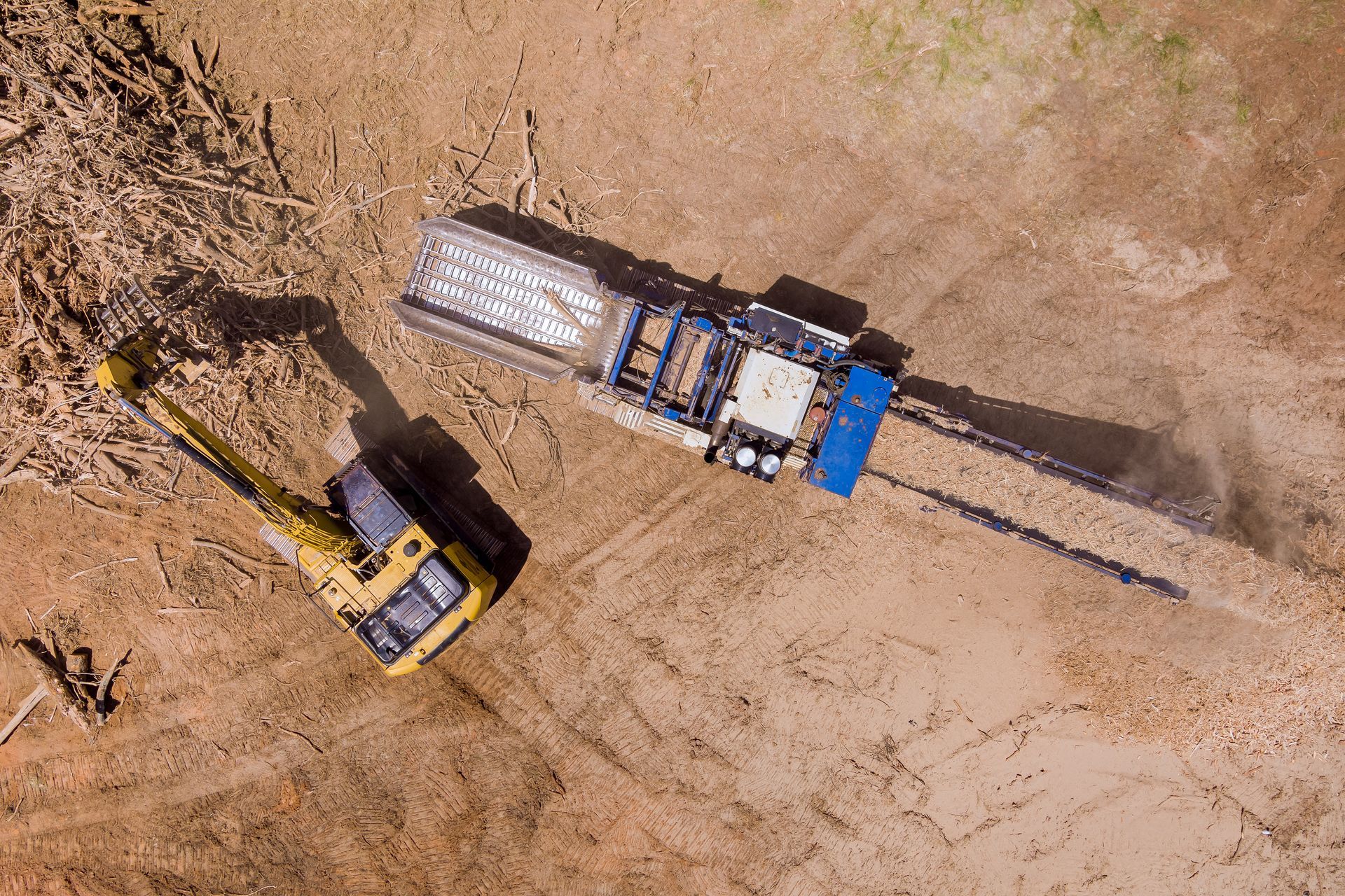 Excavator loading dirt beside an orange machine in a wooded area with autumn trees