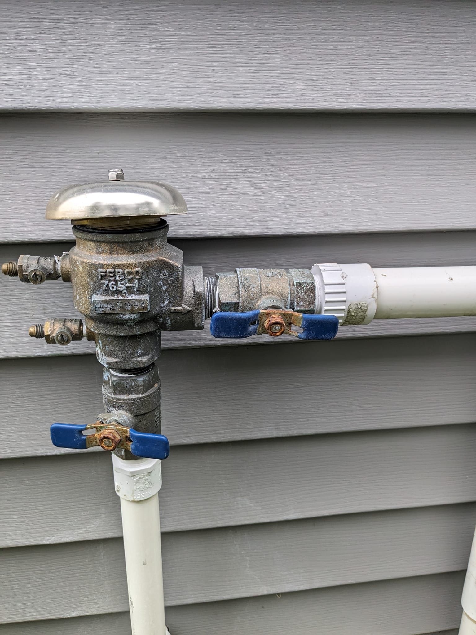 A pressure vacuum breaker irrigation assembly with two blue-handled valves mounted on a grey vinyl-sided exterior wall.
