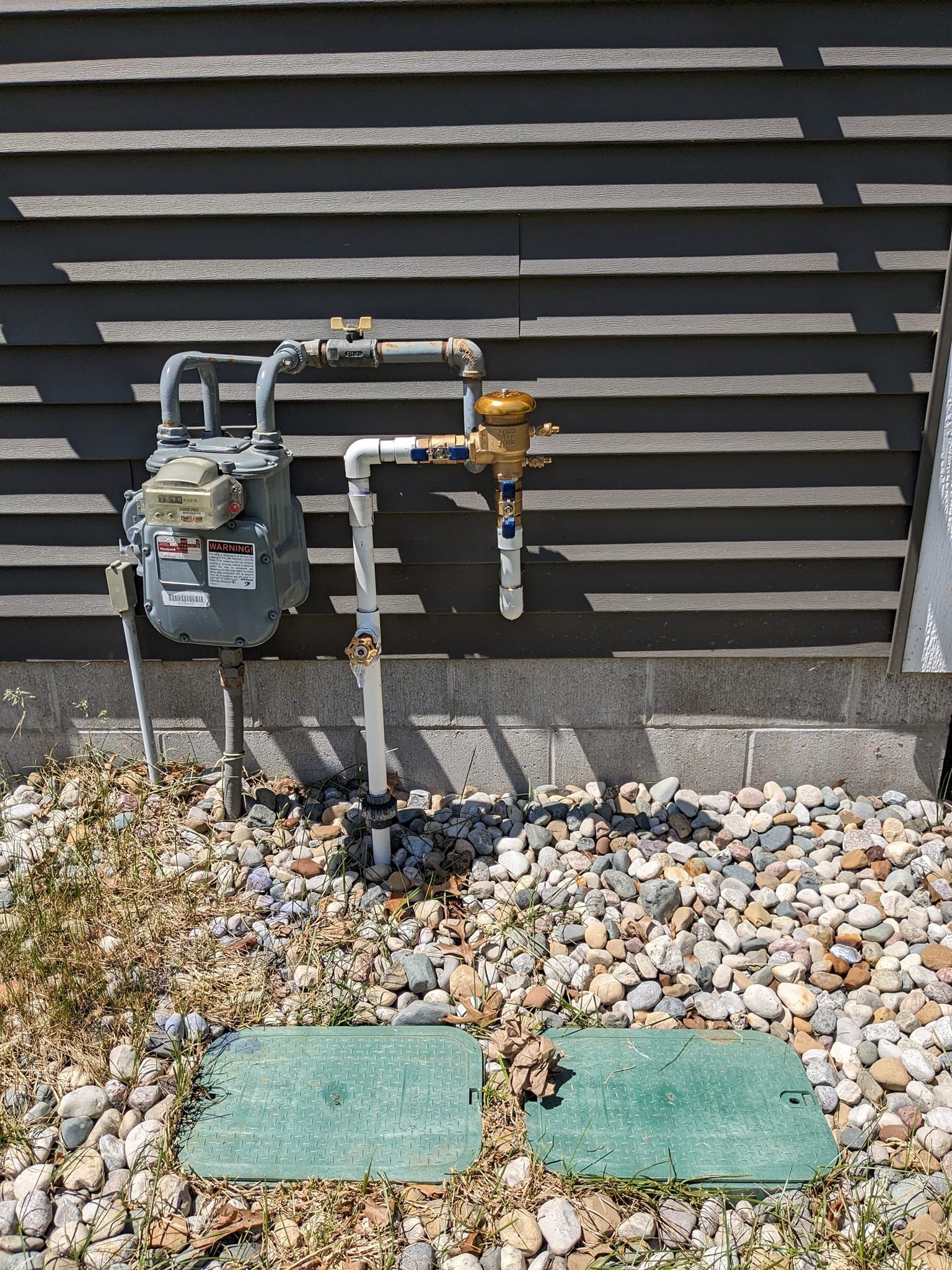 A gas meter, plumbing, and a brass valve mounted on a gray-sided wall above two green irrigation access boxes on gravel.