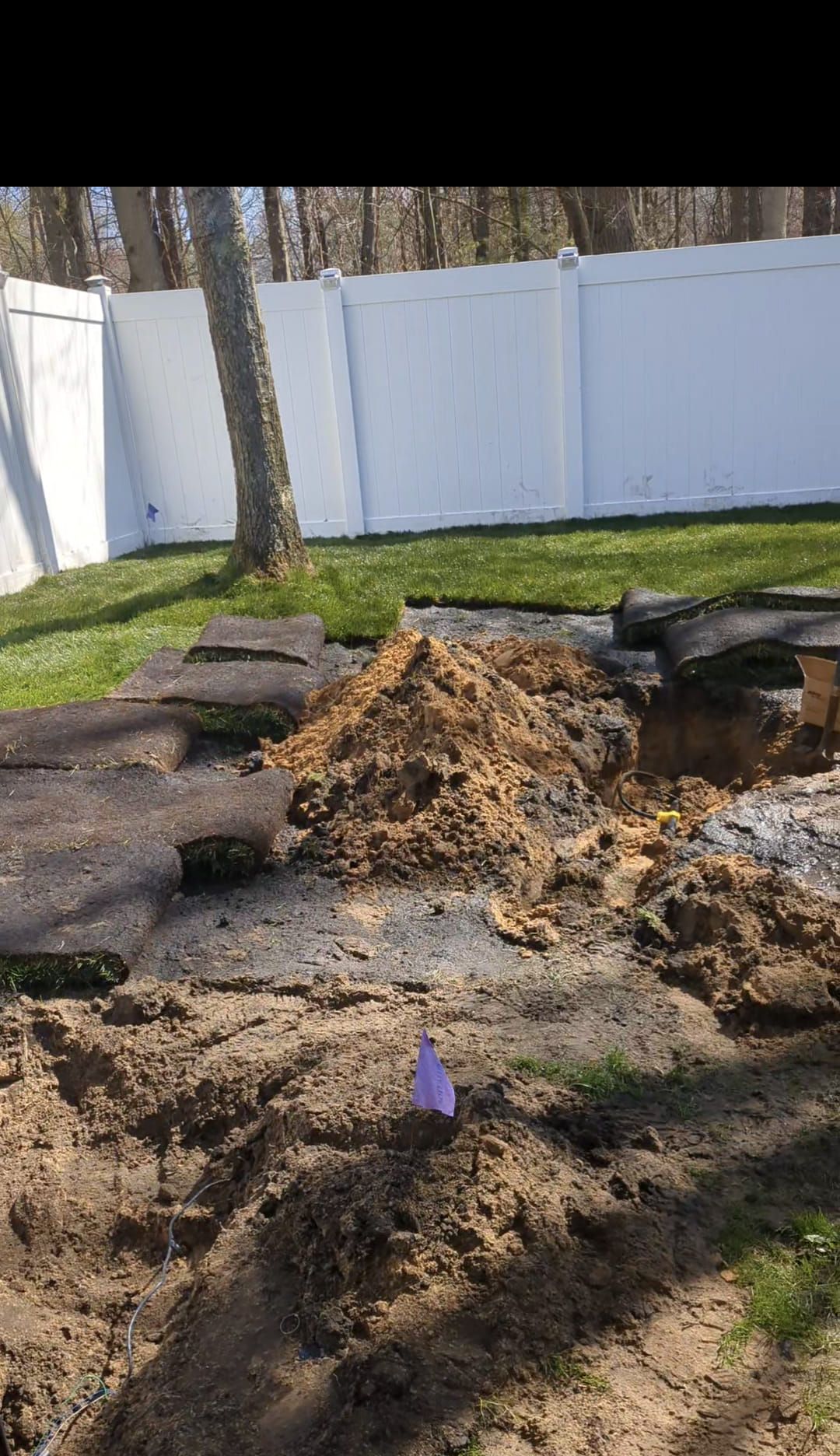 A pile of excavated dirt and pieces of sod sit in a backyard near a white privacy fence and a tree.