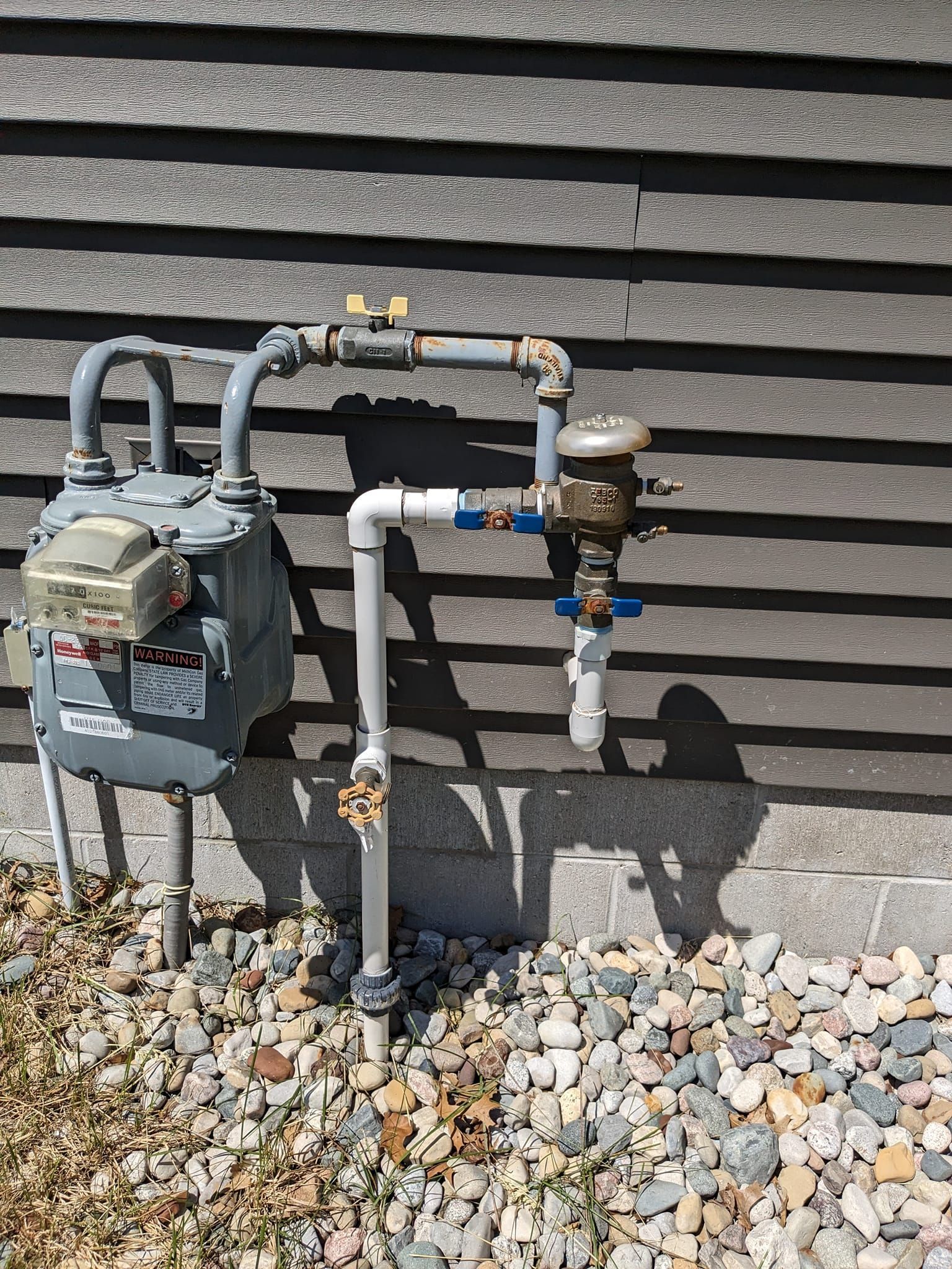 A gas meter and associated grey and white pipes mounted against a dark-sided exterior wall with gravel ground.