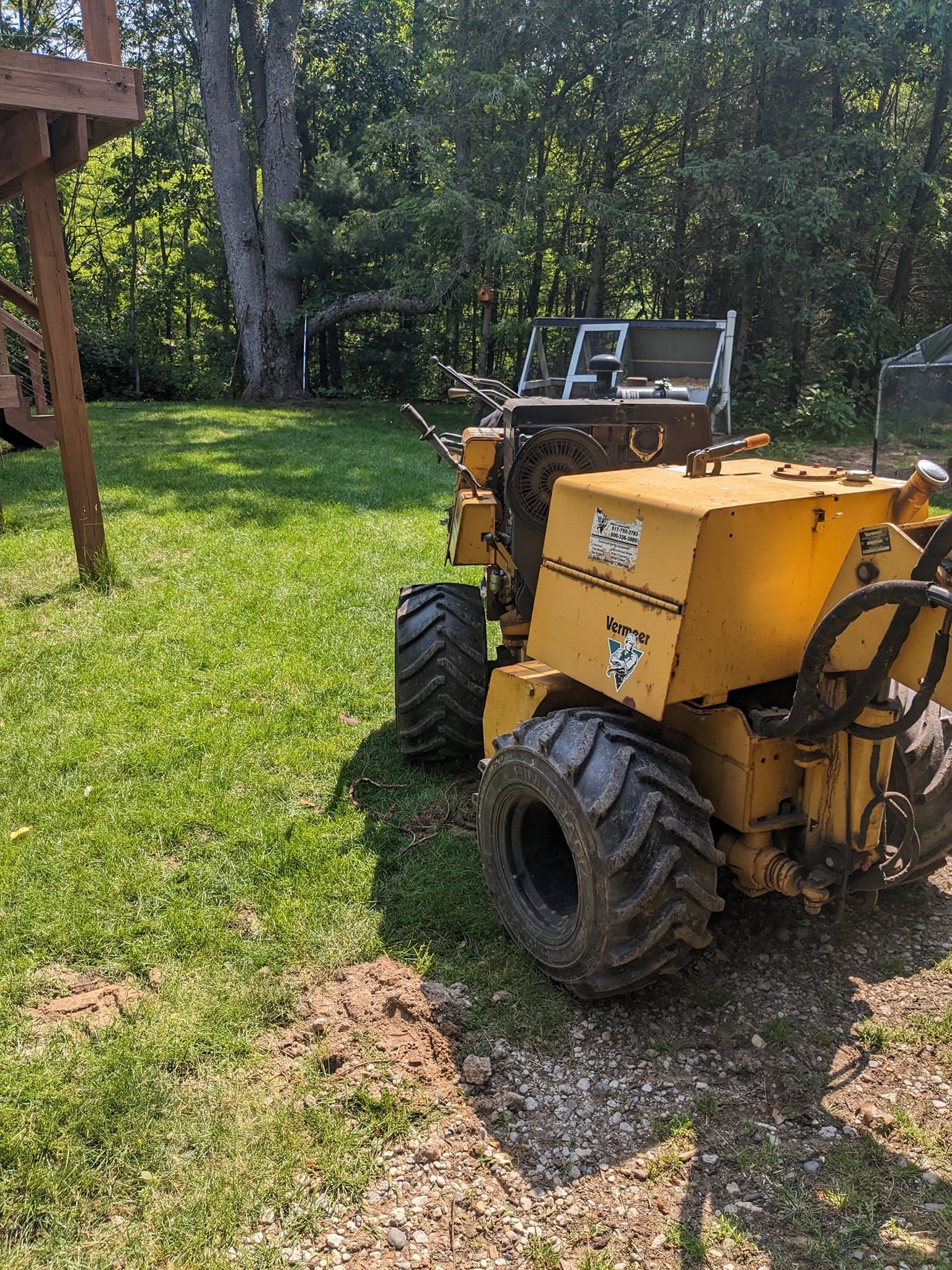 A yellow Bobcat walk-behind trencher parked on a grassy lawn near trees and a wooden deck.