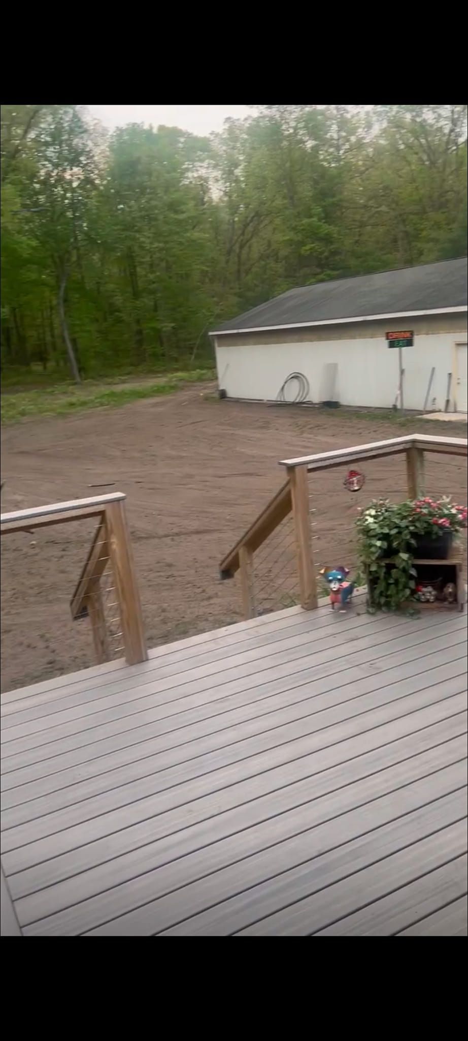 A gray wooden deck overlooks a large, empty dirt yard next to a white building, framed by a forest in the background.
