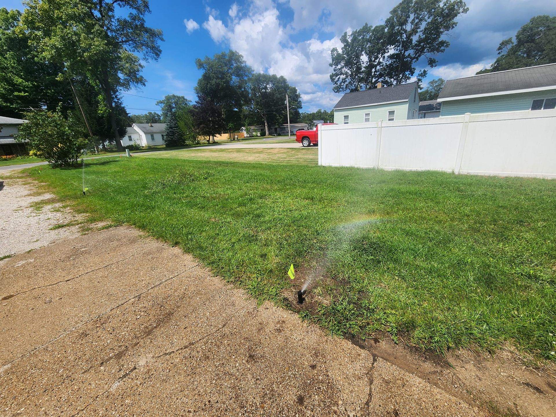 A lawn sprinkler sprays water across a green yard next to a gravel driveway, with houses and trees in the background.