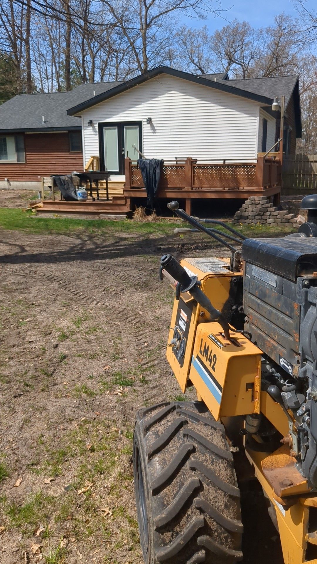 A yellow stump grinder sits on a dirt yard in front of a white house with a brown wood deck.