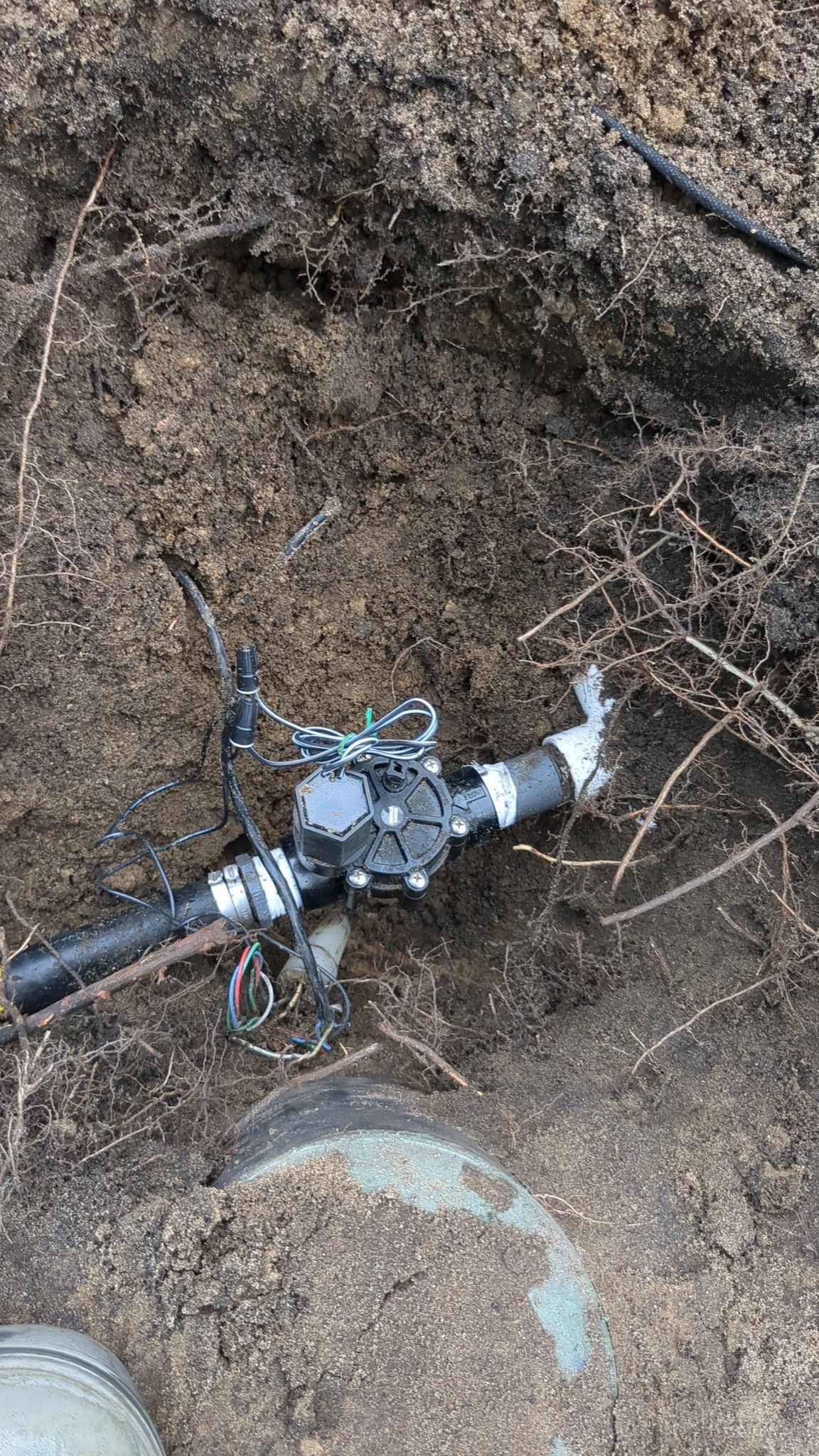 An irrigation solenoid valve connected to black plastic piping, partially uncovered in a hole filled with dirt.