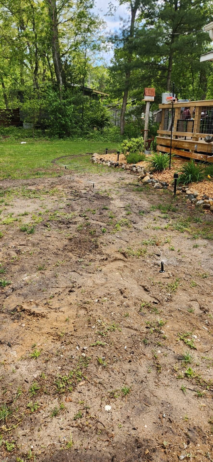 A dirt-covered yard with a landscaped edge featuring small plants, mulch, and solar lights beside a wooden deck.