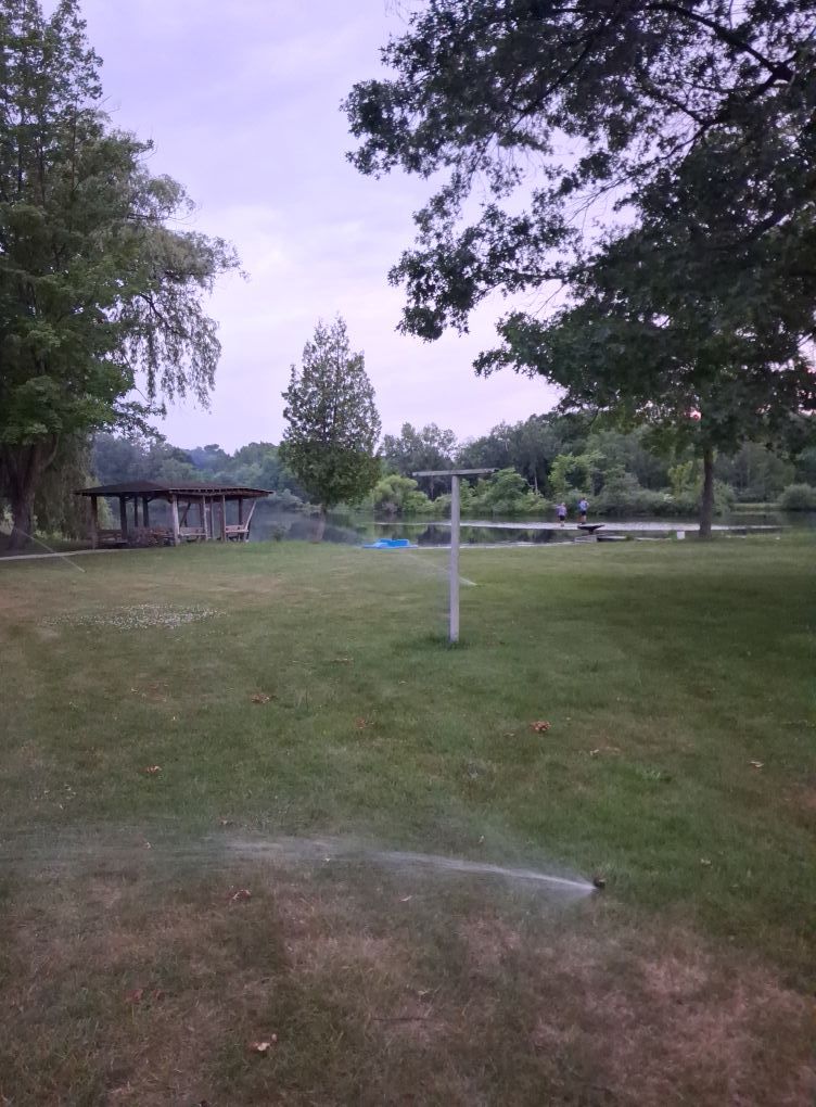 A backyard lawn with a clothesline and an active sprinkler, facing a calm lake and a small pavilion at dusk.