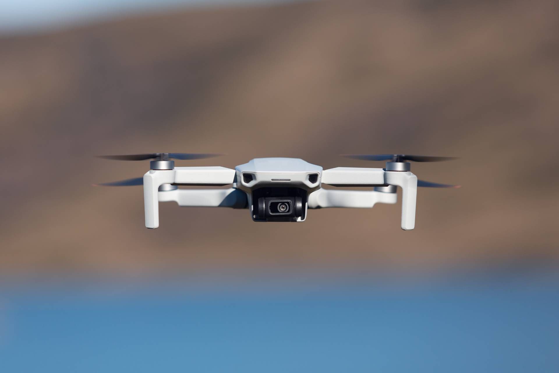 White drone flying above blue water with a blurred brown shoreline in the background