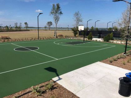 Outdoor basketball court with green surface, hoops, and a sidewalk beside a landscaped field.
