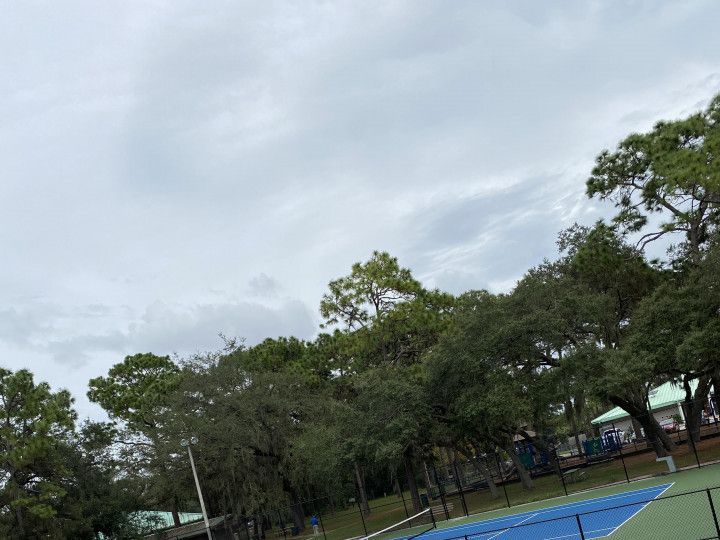 Outdoor tennis court under a cloudy sky, surrounded by trees and a blue fence