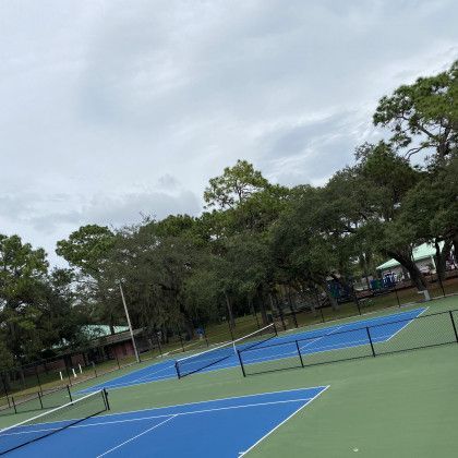 Empty outdoor tennis courts with blue and green surfaces, surrounded by trees under an overcast sky