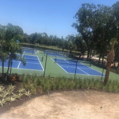 Blue tennis courts in a sunny park, surrounded by trees and a dirt path in the foreground