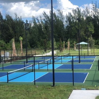 Blue and green outdoor pickleball courts with nets, surrounded by trees and grass under a partly cloudy sky