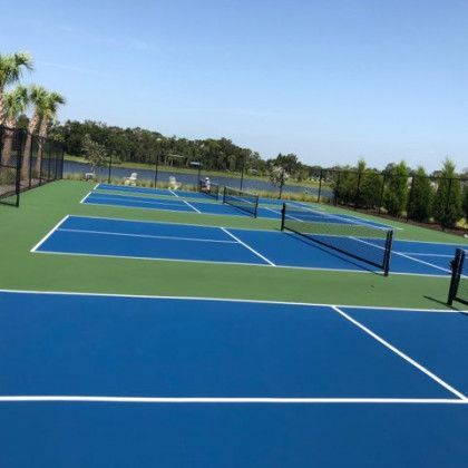 Blue tennis courts with black nets on a sunny day, surrounded by trees and a fence.