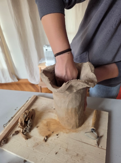 Person formt Ton in einer papierähnlichen Form, Holzbrett mit Werkzeugen in der Nähe.