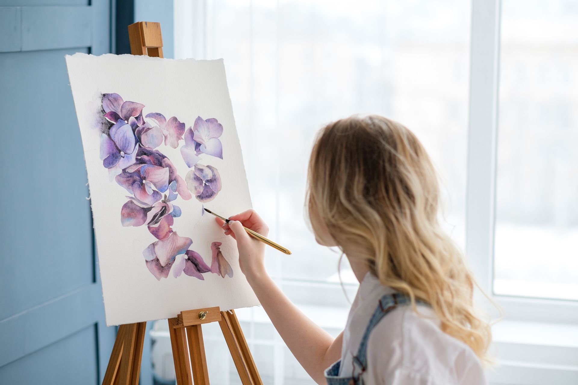 Frau mit blonden Haaren malt Hortensienblüten in Aquarell auf einer Staffelei.