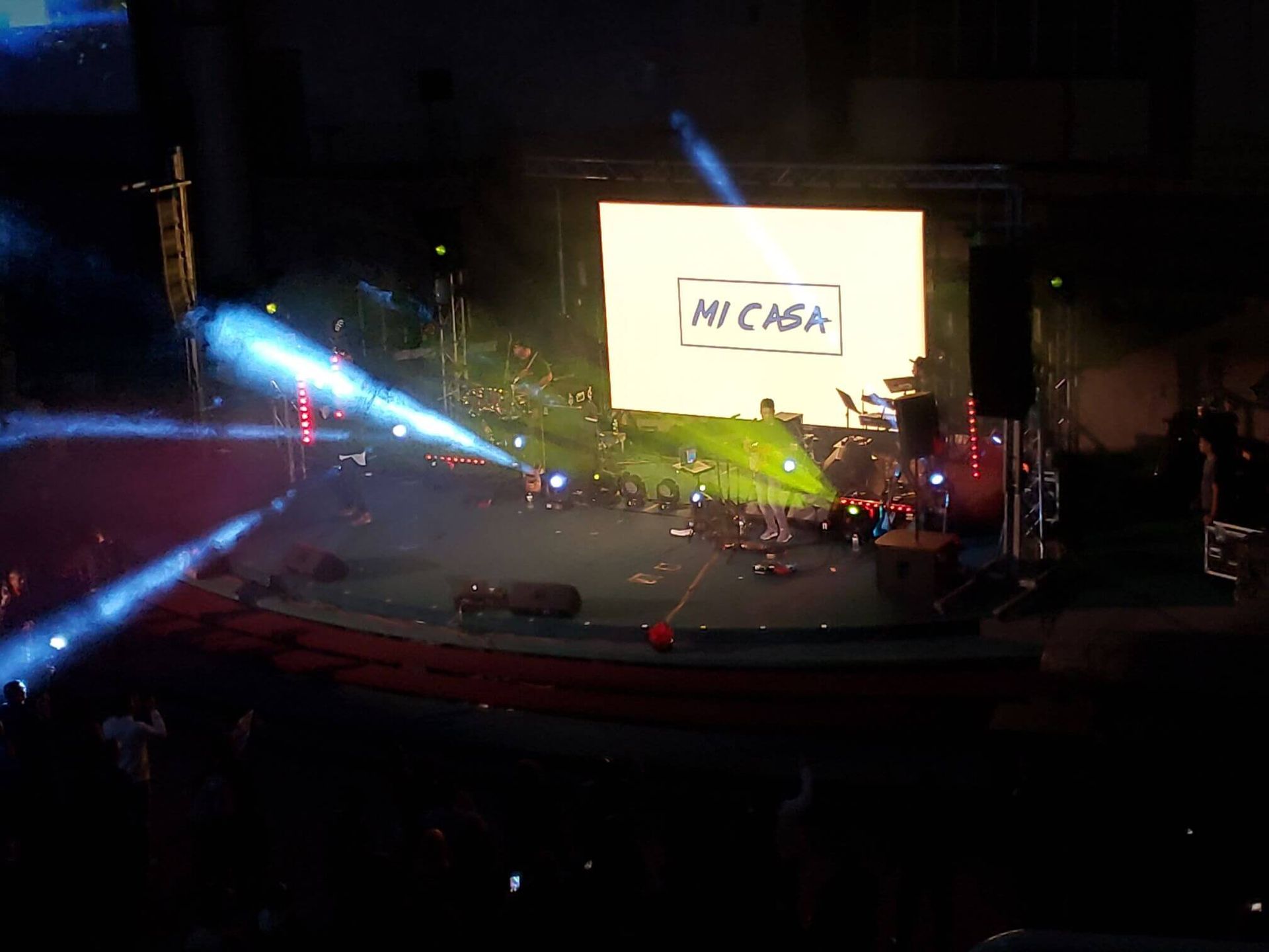 Concert stage with bright blue and red lights, crowd silhouettes, and a screen reading “MY CAFE”
