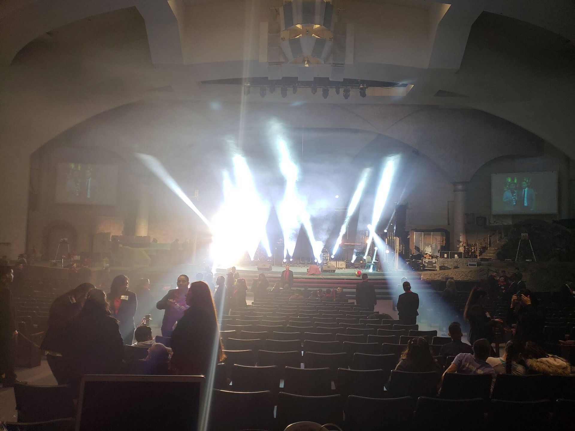 Concert hall with bright stage lights, audience seated in rows, and people milling near the aisles.
