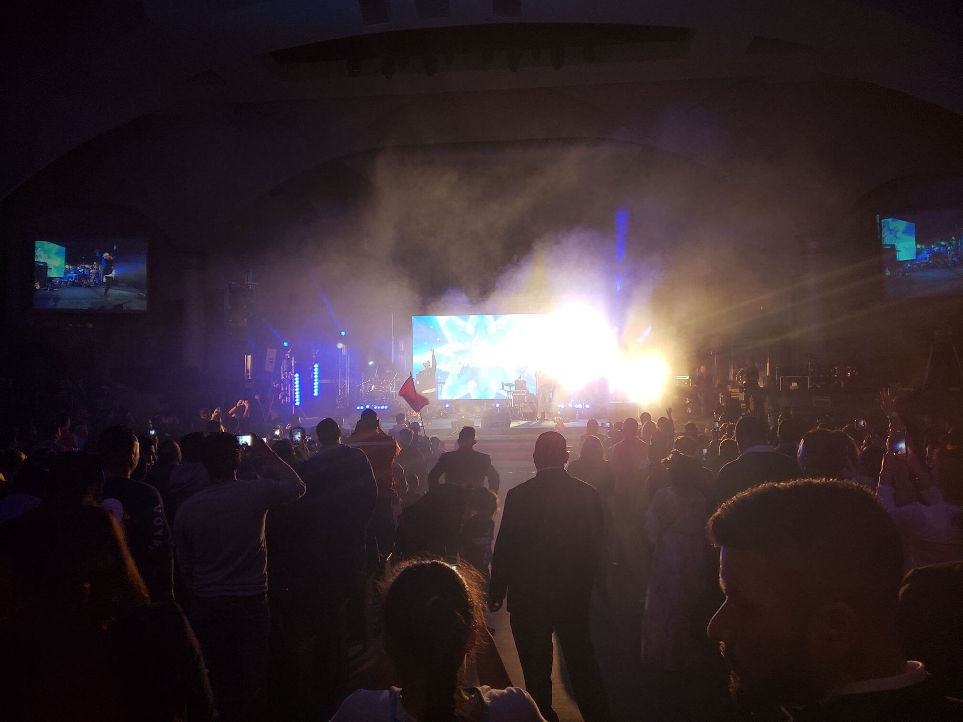 Crowded concert venue with bright stage lights and a large screen at center stage