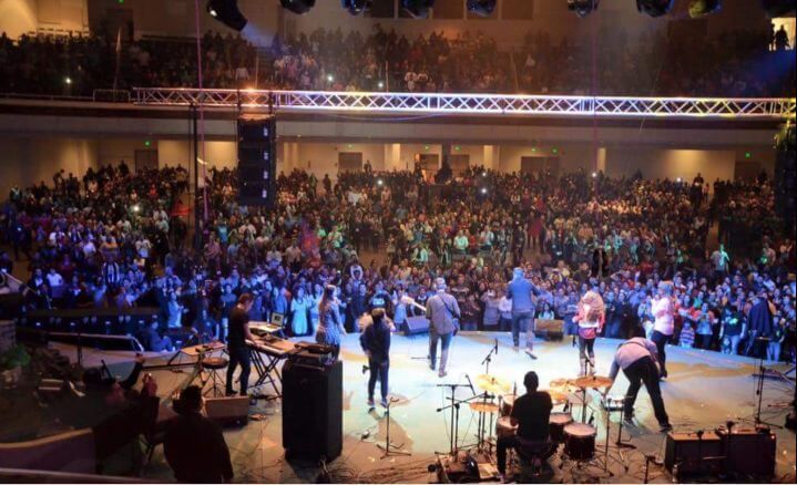 Concert stage viewed from behind performers, with a large audience in a brightly lit auditorium.
