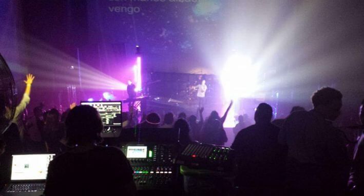 Silhouetted crowd at a concert, facing bright purple and white stage lights with DJ equipment in foreground