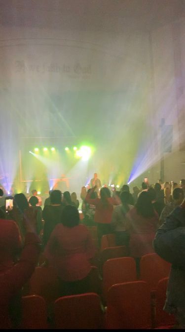 Audience seated in a dim concert hall facing bright yellow and purple stage lights