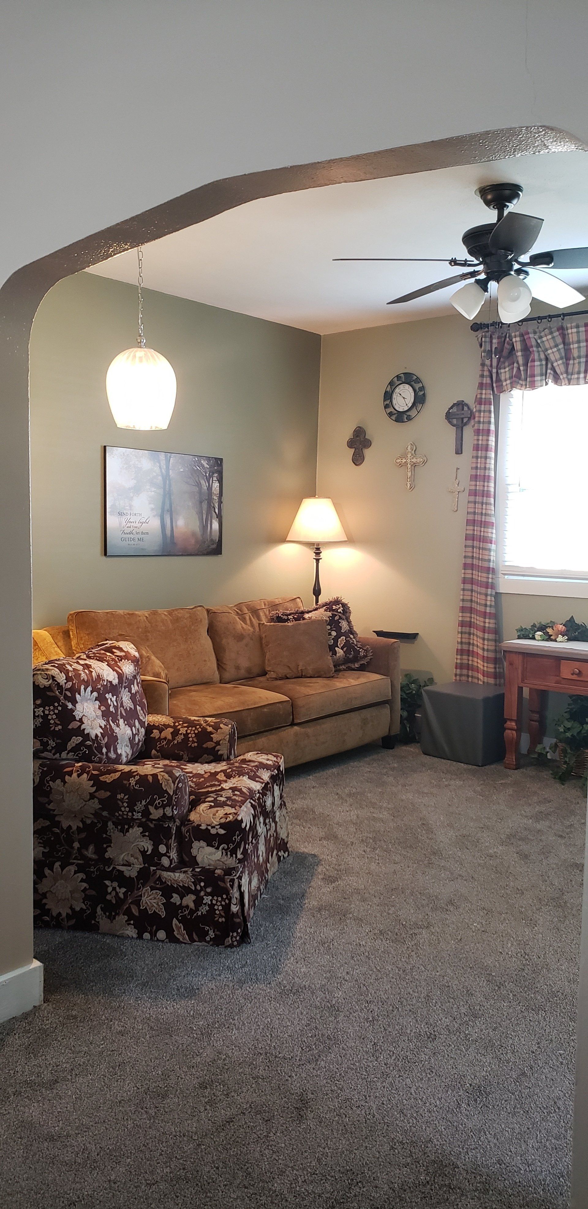 Living room with brown couch, patterned chair, gray carpet, and decorative crosses on the wall.