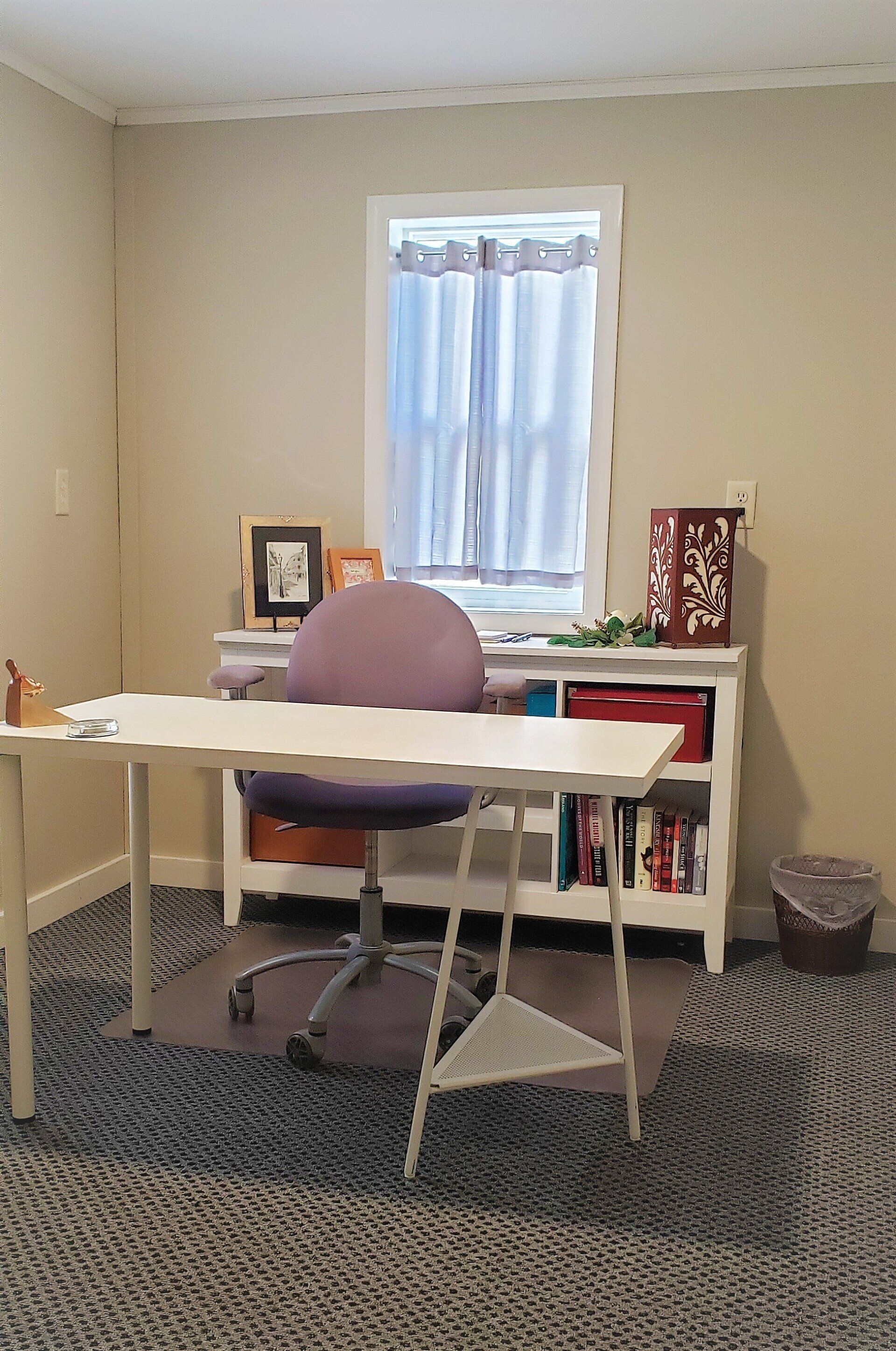 Home office with desk, purple chair, bookshelf, and window with curtain. Beige walls, patterned carpet.