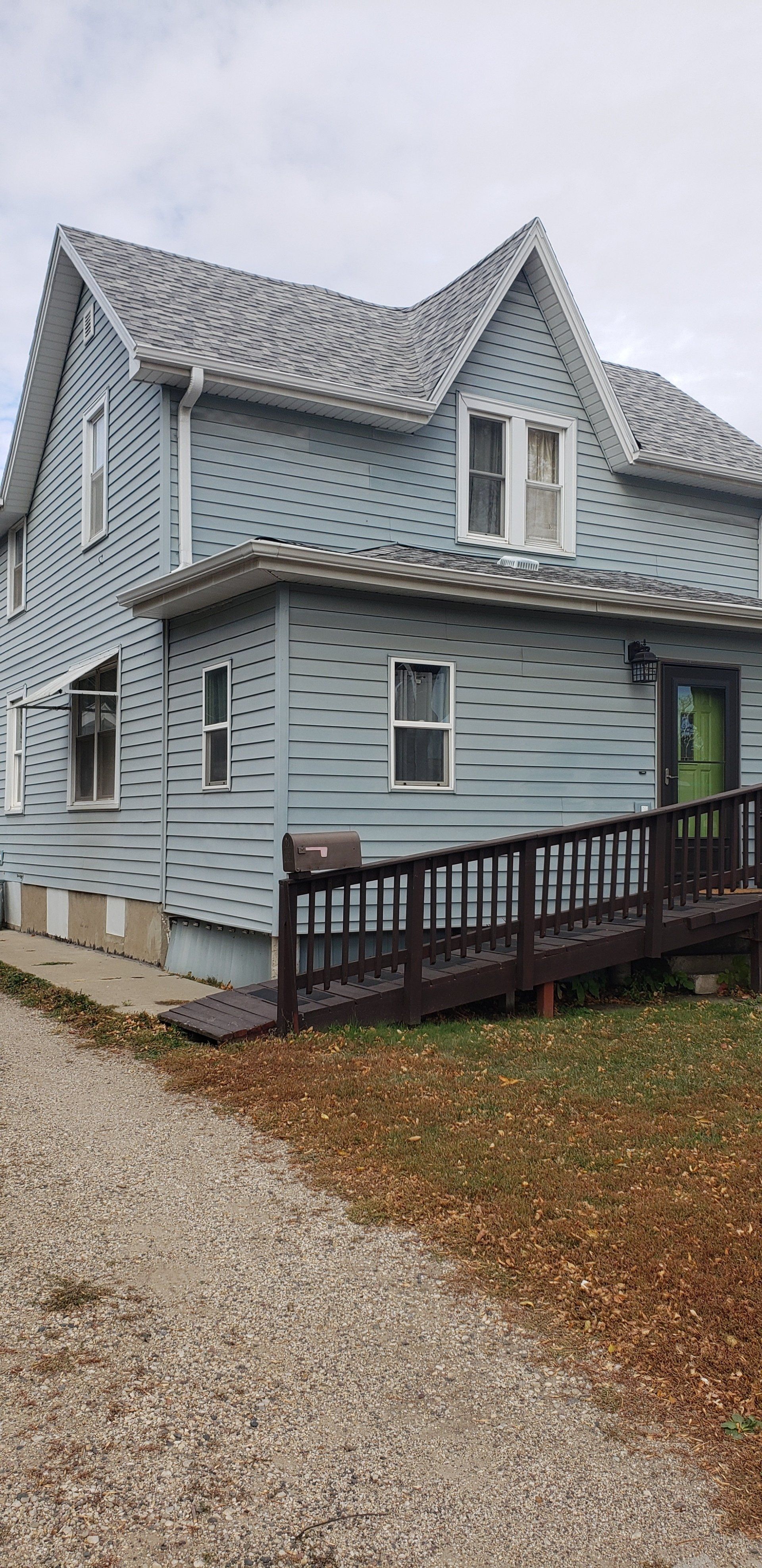 Blue house with decorative lines, ramp, and gravel driveway. Overcast sky.