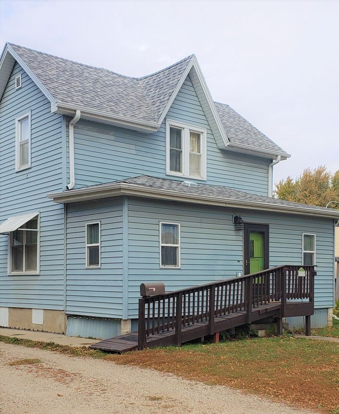 Two-story blue house with a ramp leading to a green door; gravel driveway.