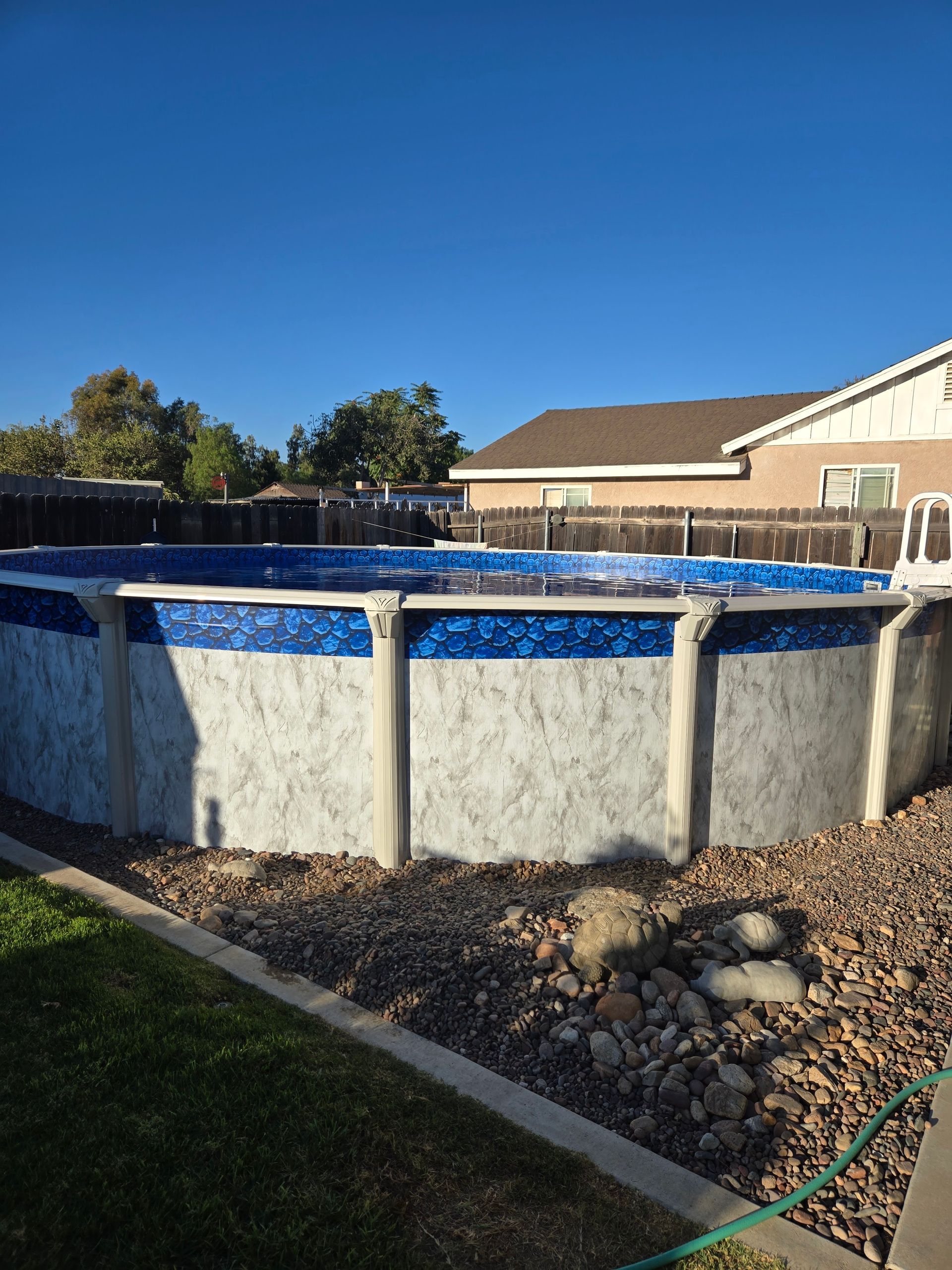 Round above-ground pool in a backyard with blue liner, fence, grass, and a clear blue sky.