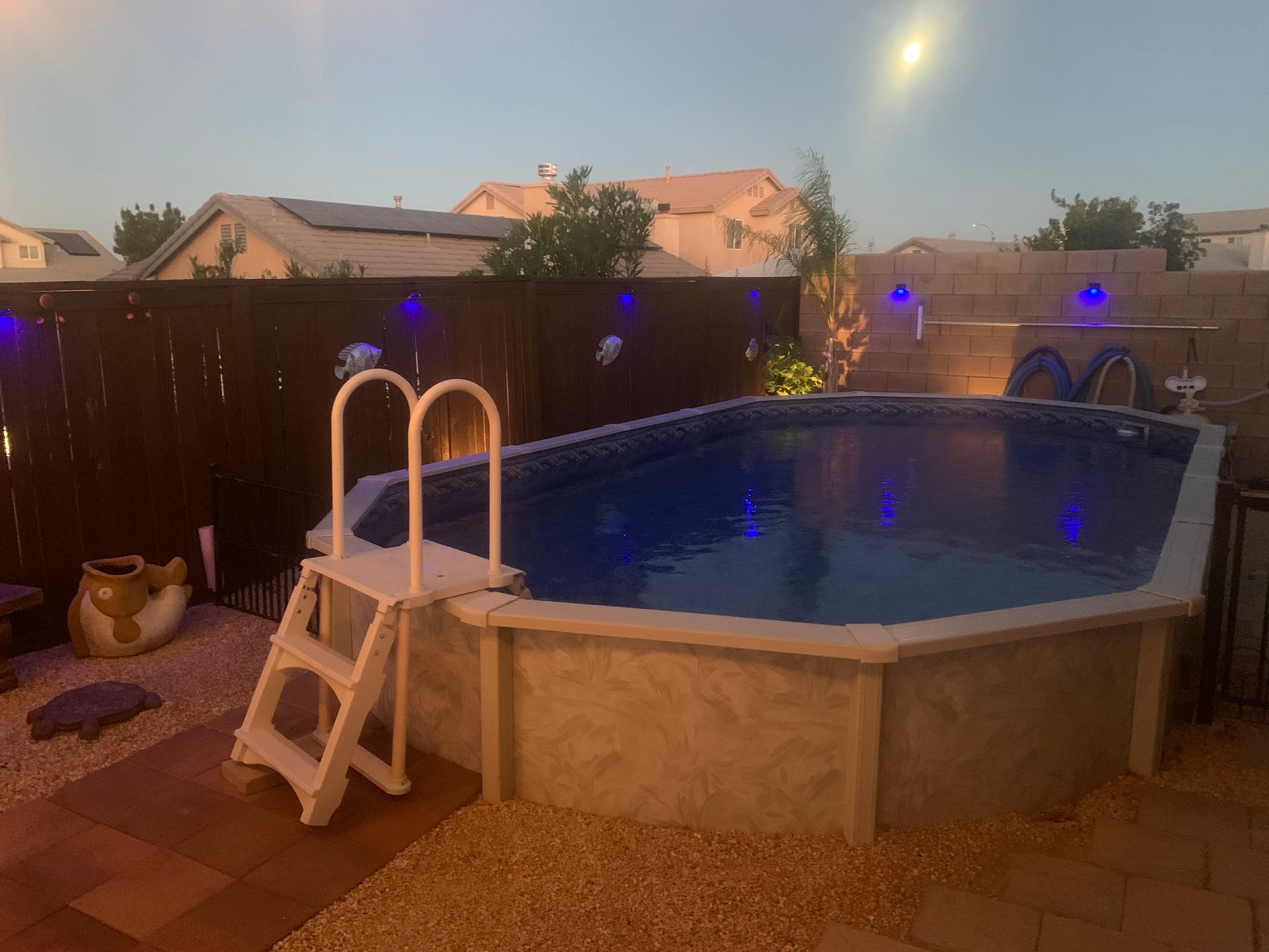 Backyard above-ground pool at dusk with blue lights, patio pavers, and a white pool ladder