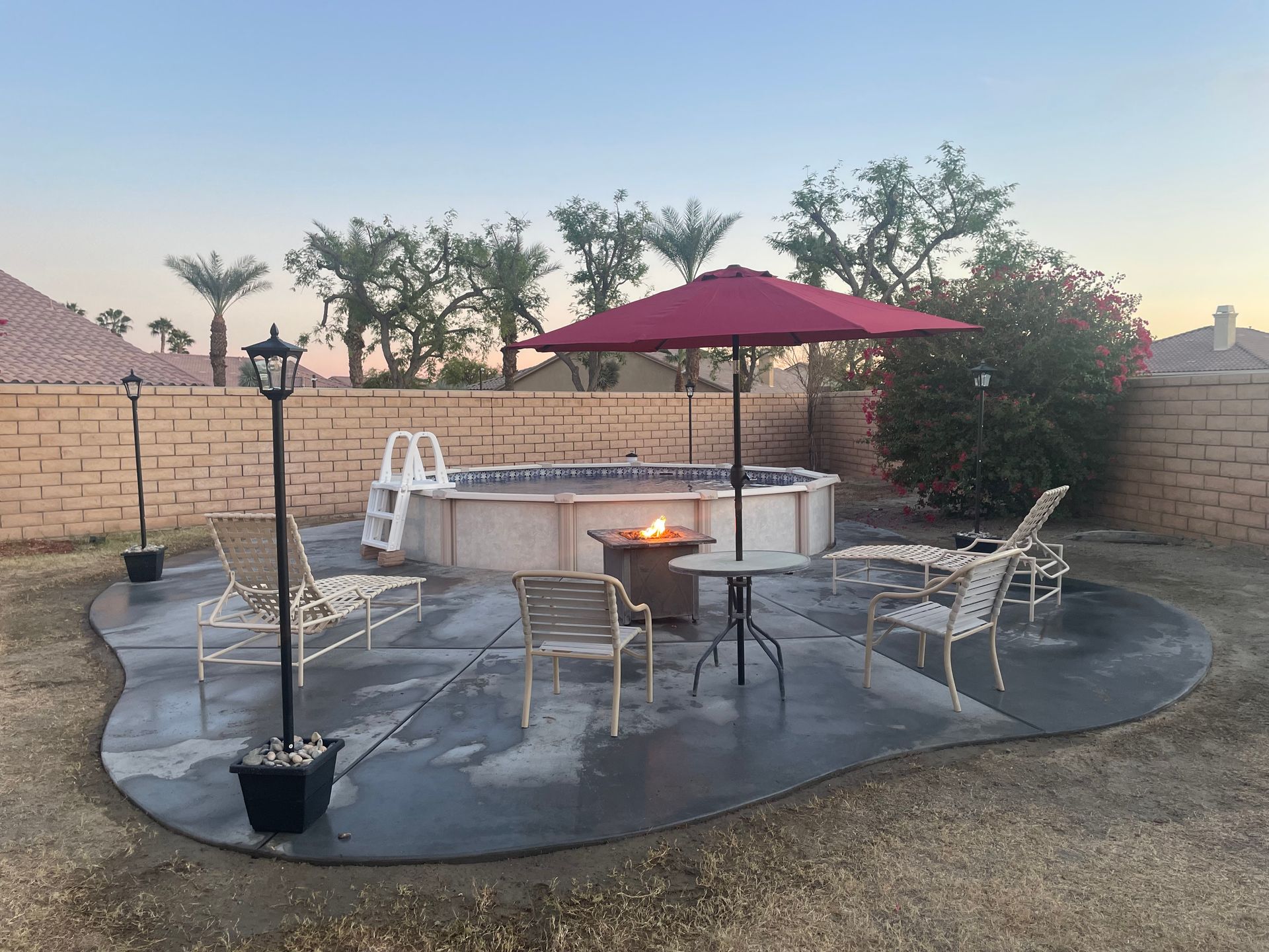 Backyard patio with a hot tub, fire pit, chairs, and a red umbrella at sunset