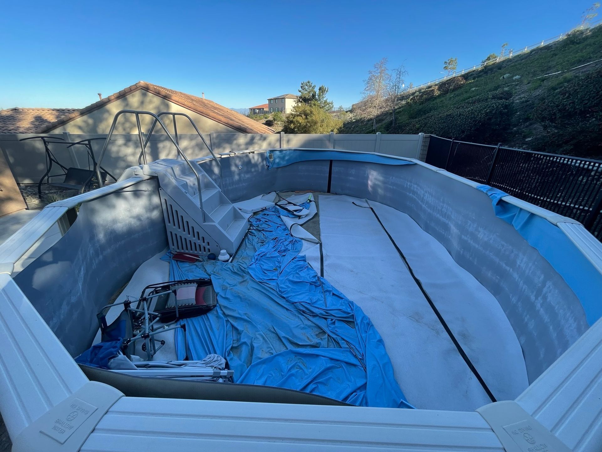 Empty above-ground pool with a blue tarp and debris, viewed in a backyard under clear sky