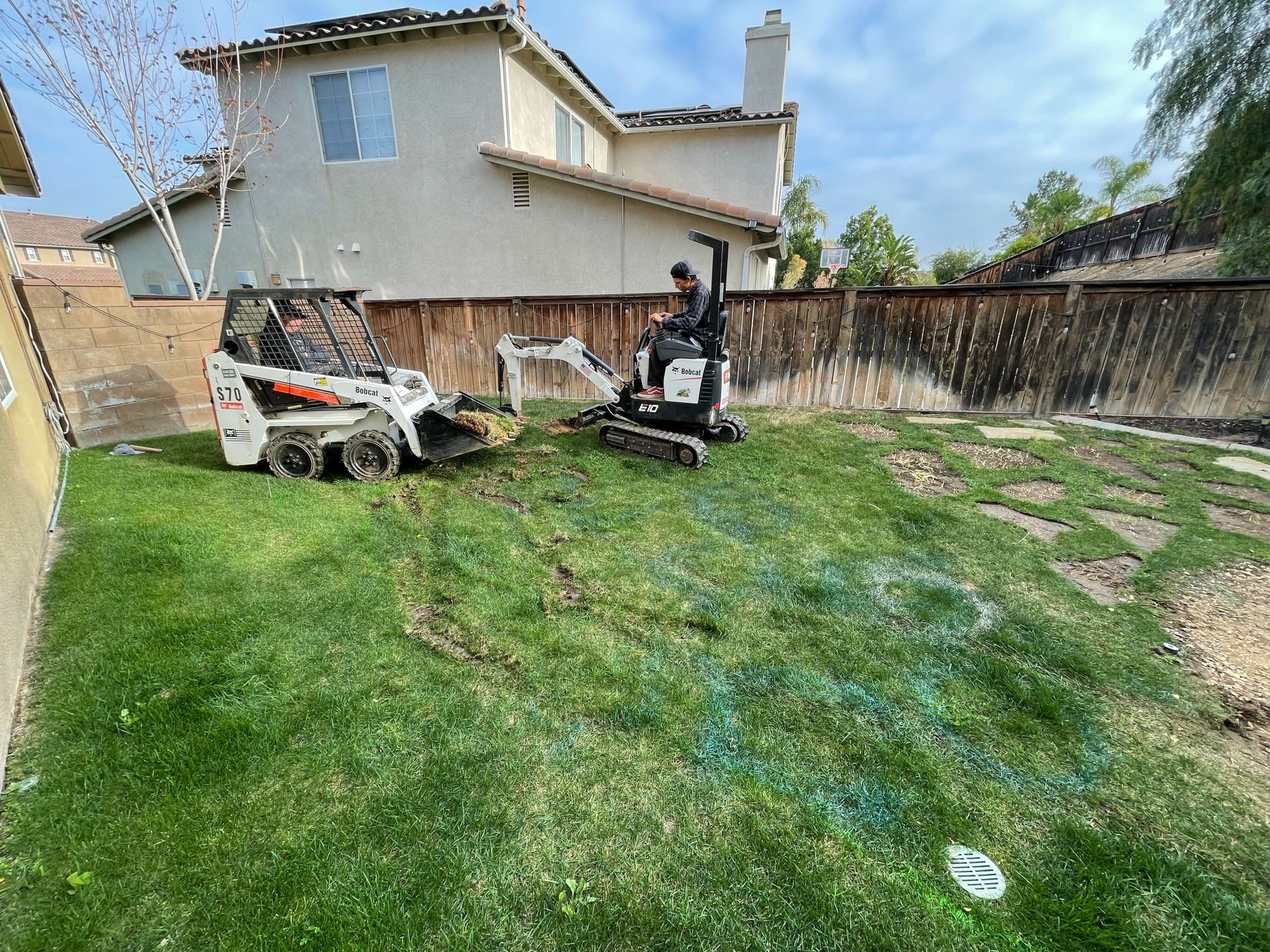 Backyard with grass, stepping stones, and two Bobcat machines near a house and wooden fence