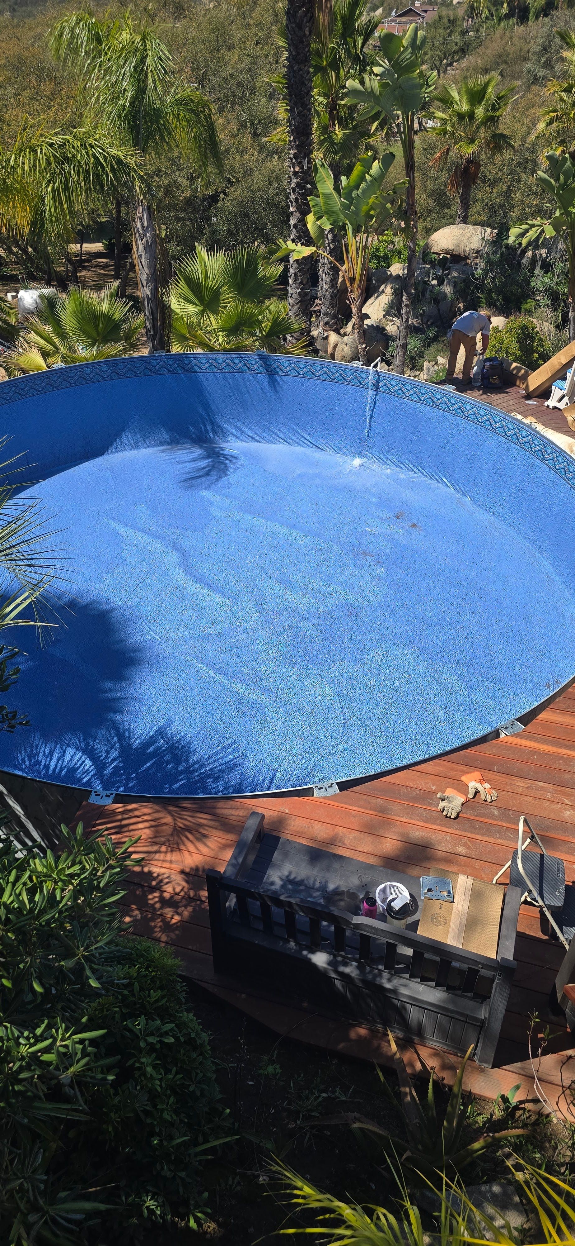 Blue round pool or pond under trees, surrounded by greenery and a brick patio.
