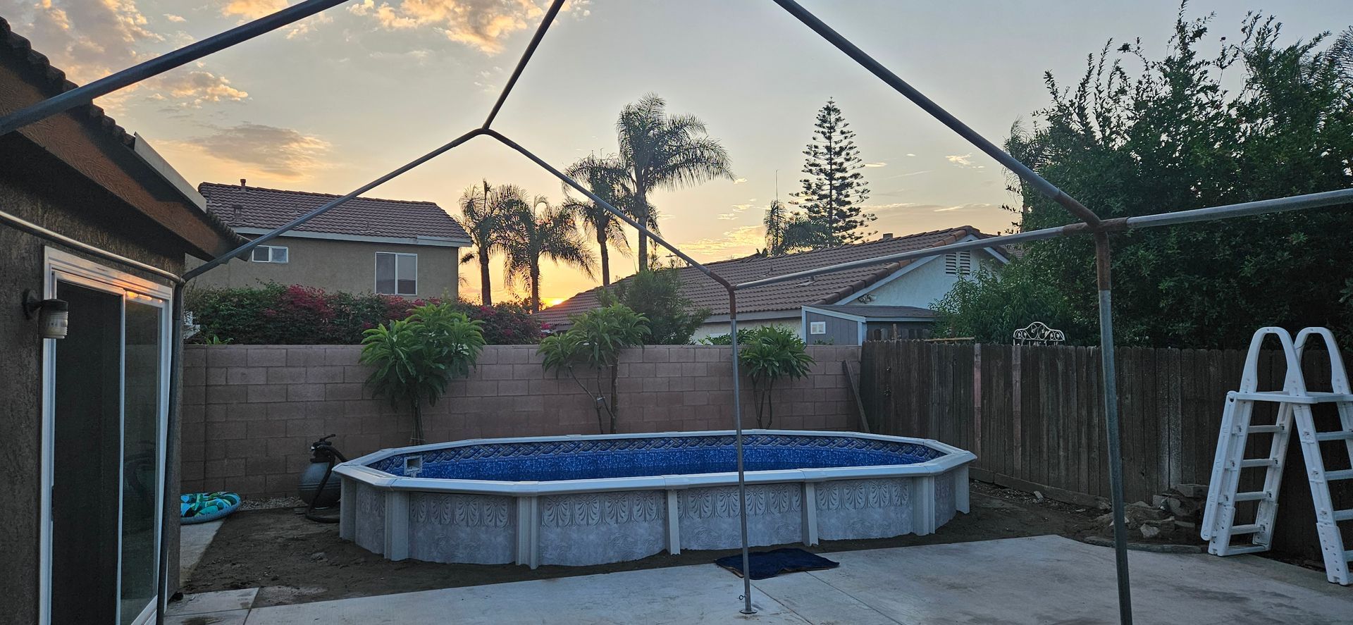 Backyard with an above-ground pool, ladder, patio, fence, and sunset sky
