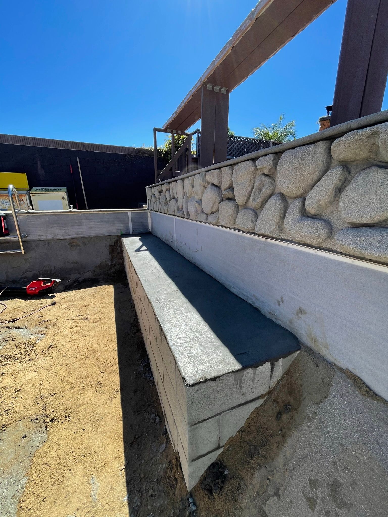 Outdoor concrete bench beside a stone retaining wall and wooden pergola in a sunny courtyard